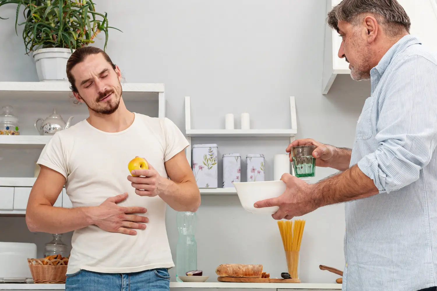 A young man stands in a kitchen holding a yellow apple, grimacing in pain with his eyes closed and one hand on his kidney, while an older man stands opposite him offering a glass of water and a white bowl